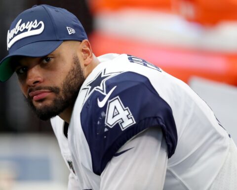 A football player in a Cowboys uniform and cap looks towards the camera while sitting on the sideline.