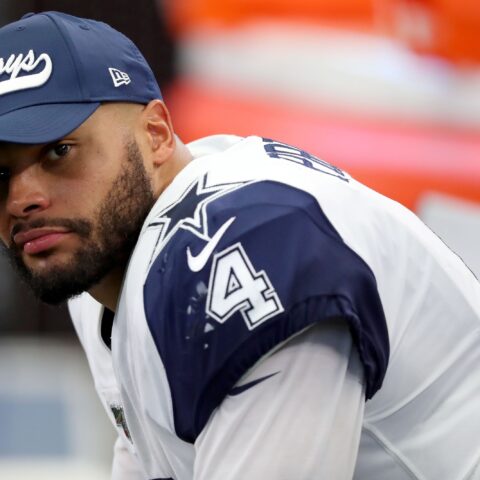 A football player in a Cowboys uniform and cap looks towards the camera while sitting on the sideline.