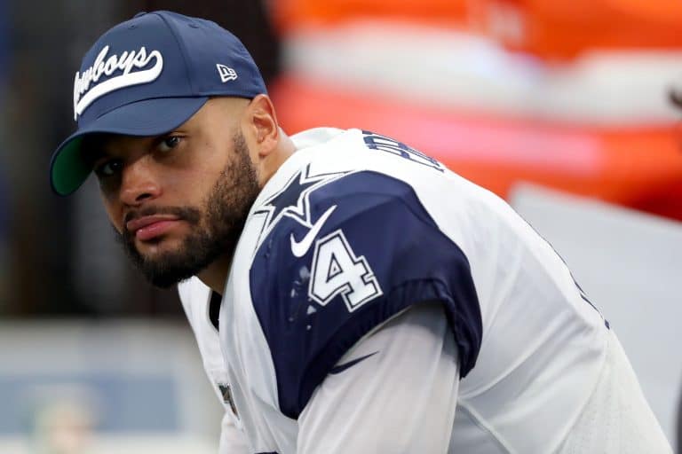 A football player in a Cowboys uniform and cap looks towards the camera while sitting on the sideline.