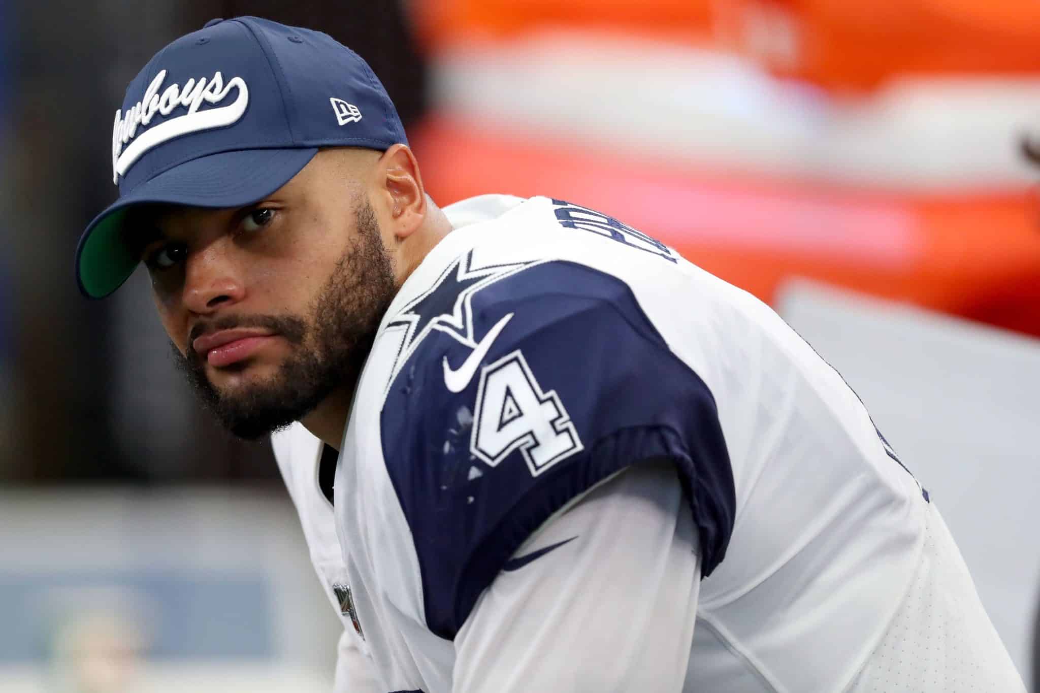 A football player in a Cowboys uniform and cap looks towards the camera while sitting on the sideline.