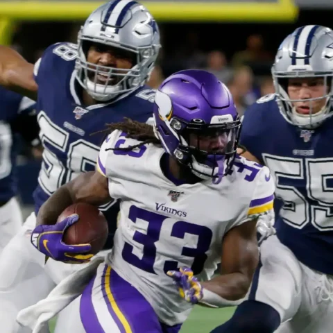 Football player in a purple jersey runs with the ball, pursued by three players in navy blue jerseys on a field.