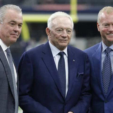 Three men in suits stand together, smiling, in a stadium setting.