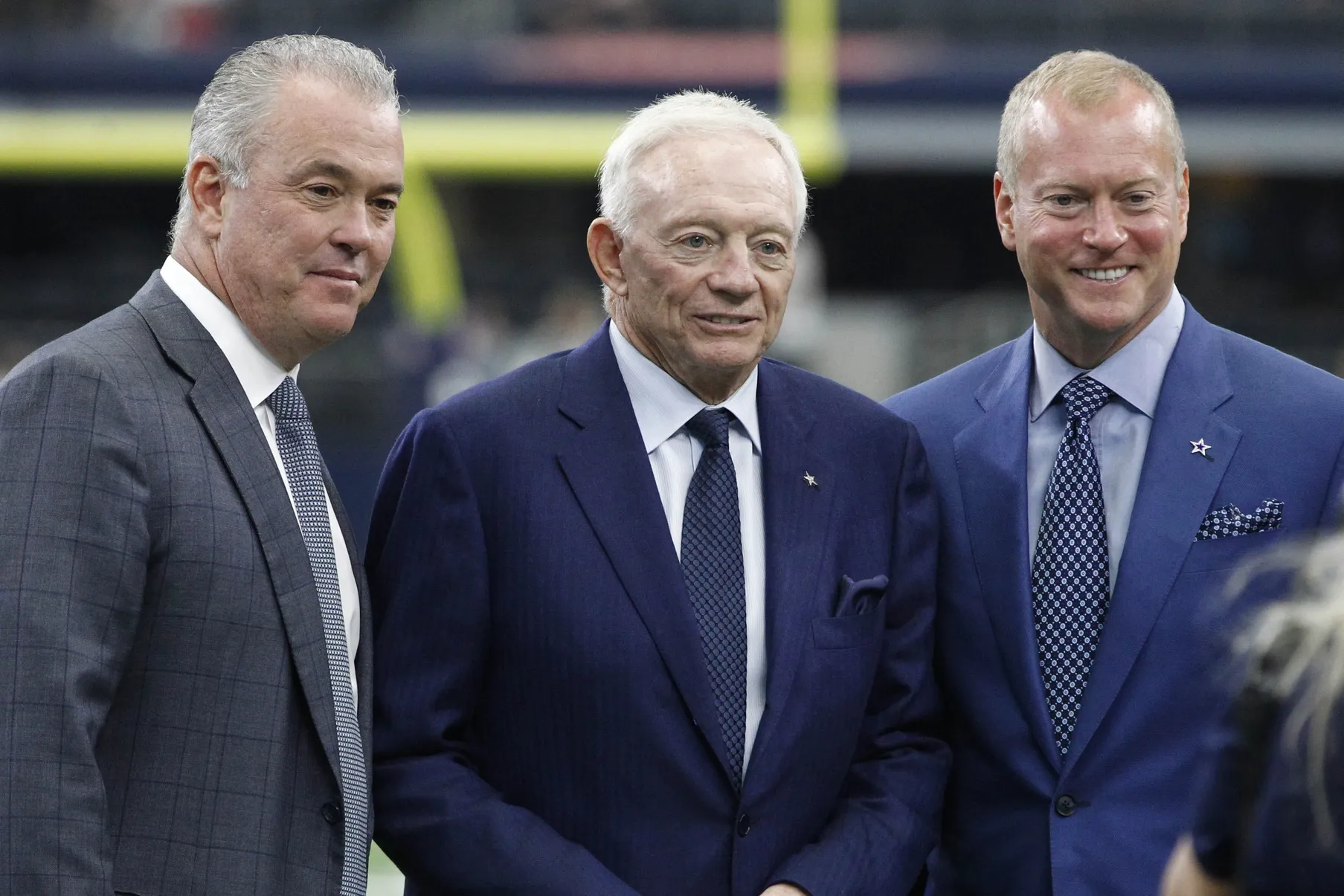 Three men in suits stand together, smiling, in a stadium setting.