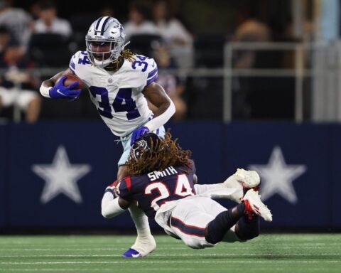 Football player in a white jersey runs with the ball while a player in a black jersey dives to tackle him.