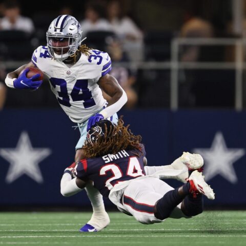 Football player in a white jersey runs with the ball while a player in a black jersey dives to tackle him.