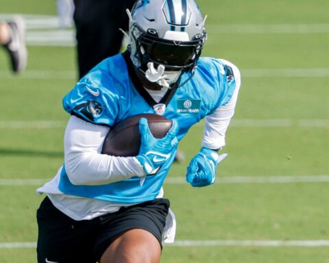 Football player in a blue jersey, holding a football, running on a grassy field during practice.