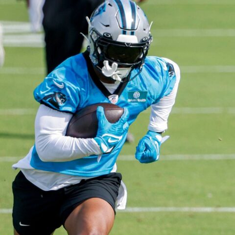Football player in a blue jersey, holding a football, running on a grassy field during practice.