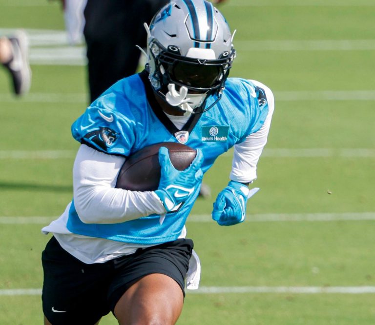 Football player in a blue jersey, holding a football, running on a grassy field during practice.