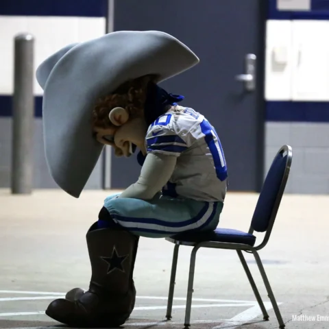 A sports mascot in cowboy attire sits on a chair with a slumped posture in a hallway.