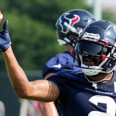 Football player in navy uniform practices throwing a football during a training session, with teammates in the background.