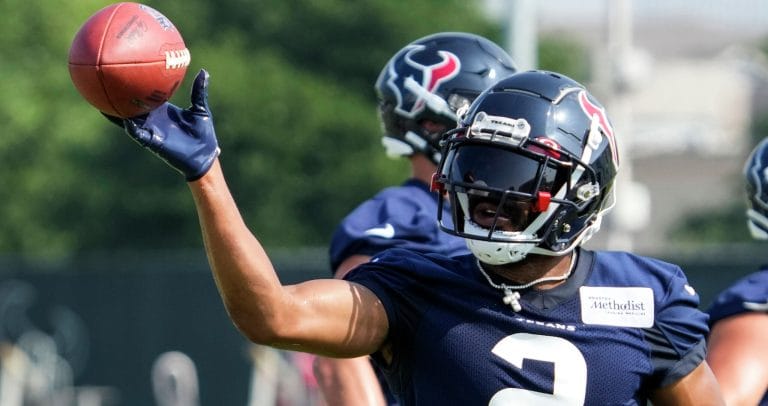 Football player in navy uniform practices throwing a football during a training session, with teammates in the background.