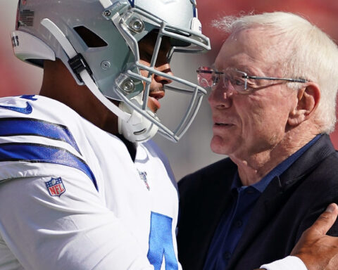 A football player in uniform and a man in a suit embrace on the field.