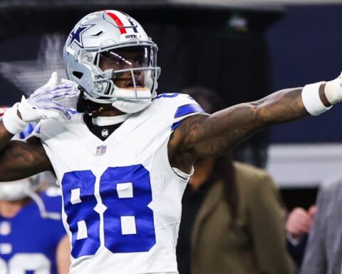 Football player in a white and blue uniform pointing while celebrating during a game. Fans and staff in the background.