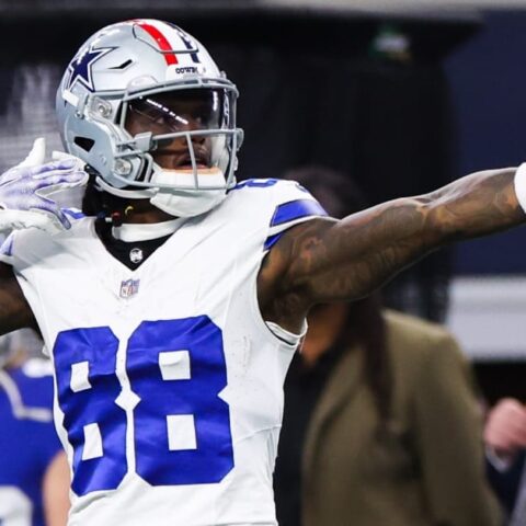Football player in a white and blue uniform pointing while celebrating during a game. Fans and staff in the background.