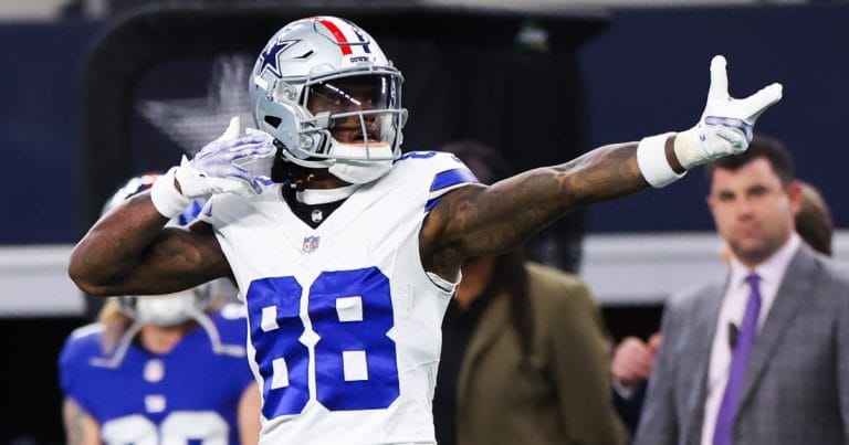 Football player in a white and blue uniform pointing while celebrating during a game. Fans and staff in the background.