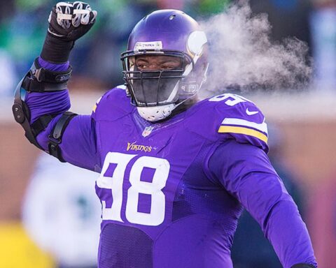 Football player in a purple jersey with number 98 raises his arm on a cold day, visible breath in the air.