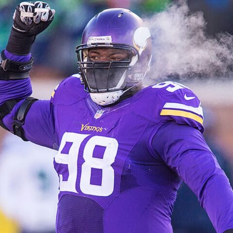 Football player in a purple jersey with number 98 raises his arm on a cold day, visible breath in the air.