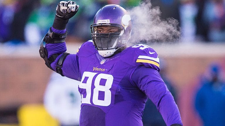 Football player in a purple jersey with number 98 raises his arm on a cold day, visible breath in the air.