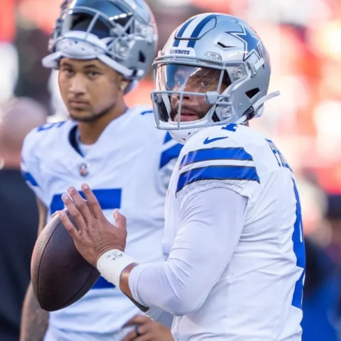 Two football players in white and blue uniforms, one holding a football, stand on the sideline during a game.