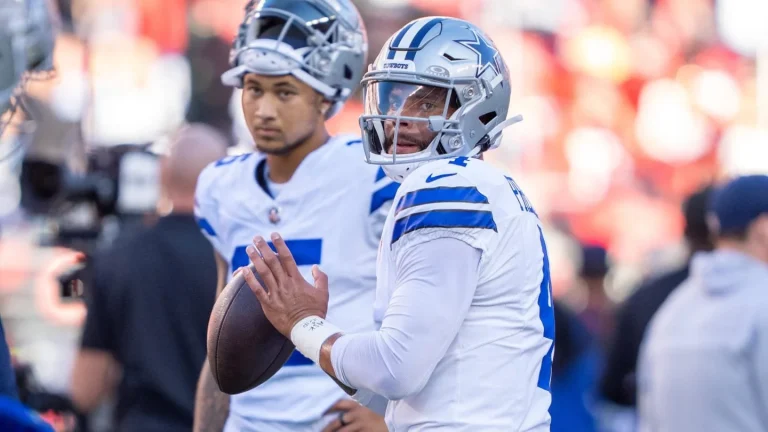 Two football players in white and blue uniforms, one holding a football, stand on the sideline during a game.