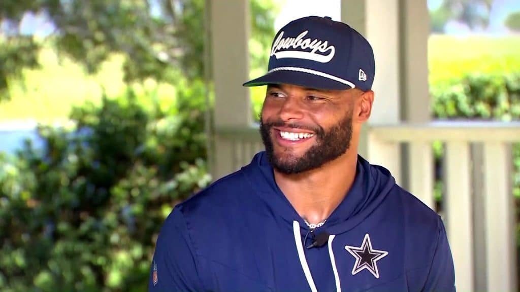 Dak Prescott's latest quote shows he's just like us - Dak Prescott, Quarterback A man in a navy blue Cowboys cap and hoodie with a logo smiles while outdoors under a covered area.