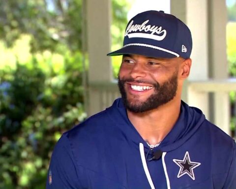 A man in a navy blue Cowboys cap and hoodie with a logo smiles while outdoors under a covered area.