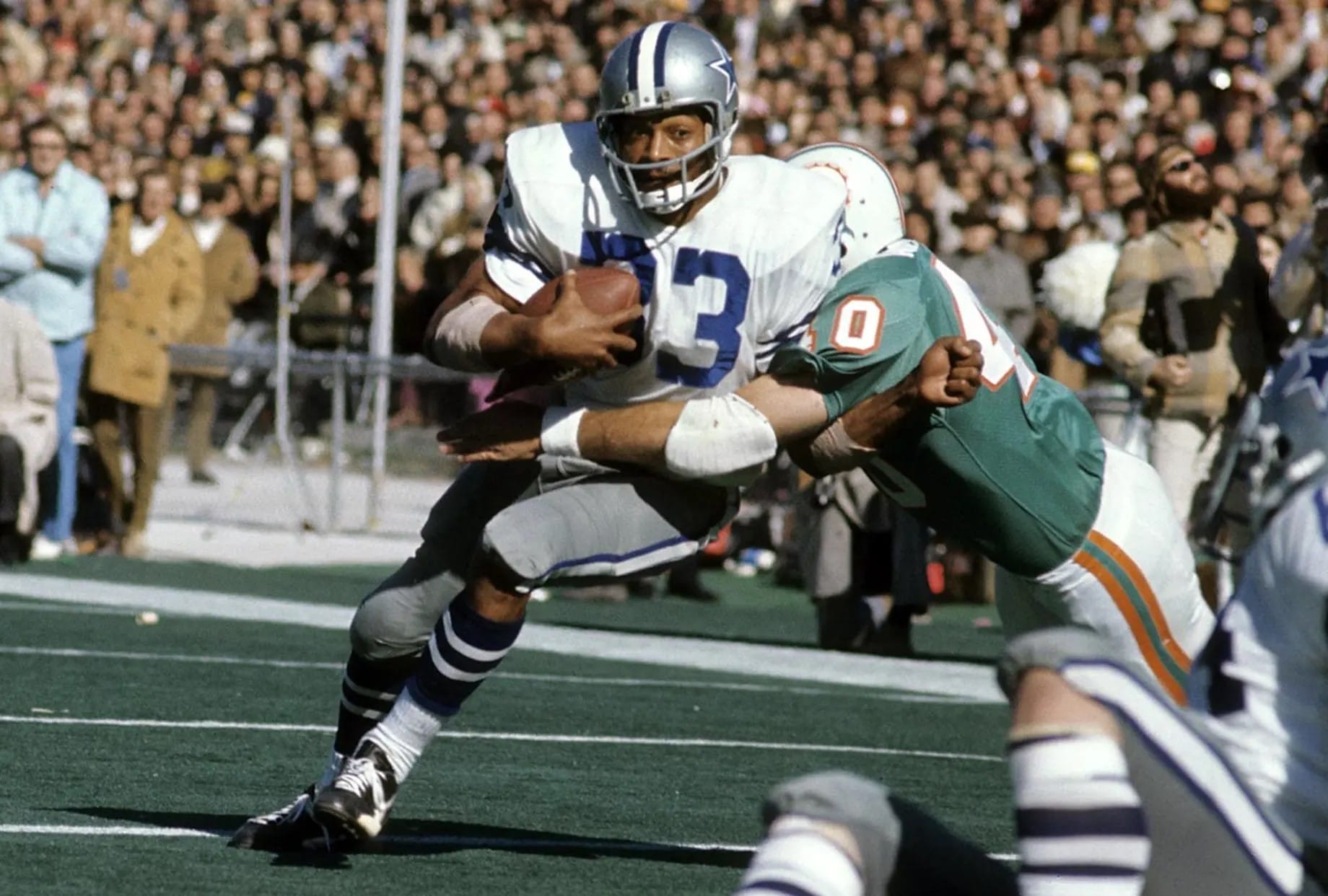 A football player in a white and blue jersey runs the ball while being tackled by a player in a white and aqua jersey.