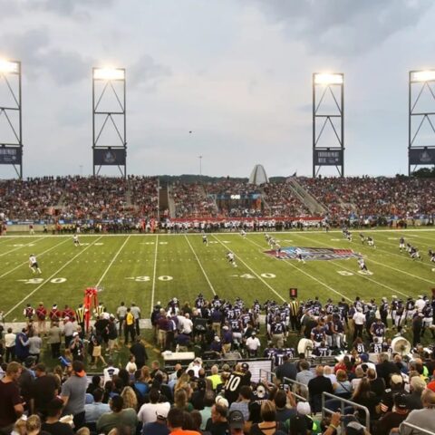 Packed football stadium with fans watching a game under bright lights during twilight, with the field in active play and incorporating recent changes to enhance the experience.