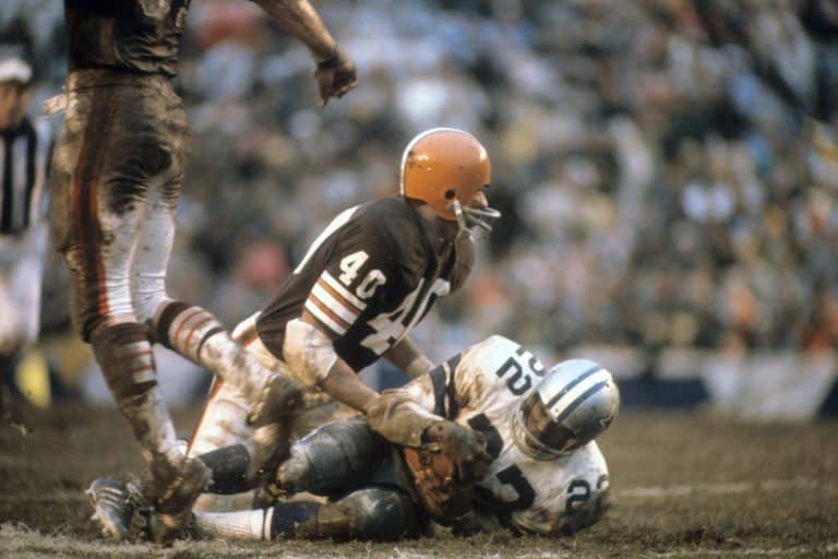 Two American football players in a tackle, one from a brown team and the other from a white and blue team, on a muddy field.