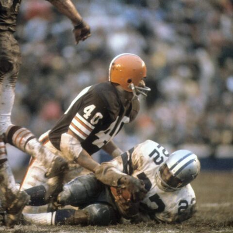 Two American football players in a tackle, one from a brown team and the other from a white and blue team, on a muddy field.