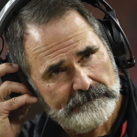 Man with a thick white beard, wearing headphones and adjusting the left earpiece.