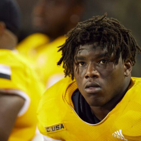 A football player wearing a yellow jersey with the text "CUSA," looking focused while on the sidelines.