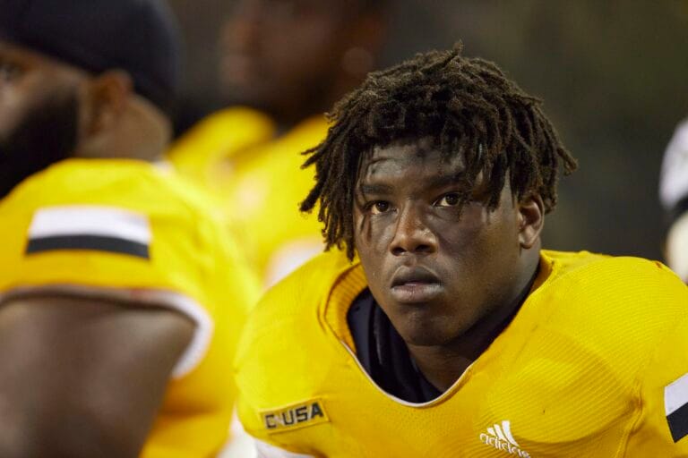 A football player wearing a yellow jersey with the text "CUSA," looking focused while on the sidelines.