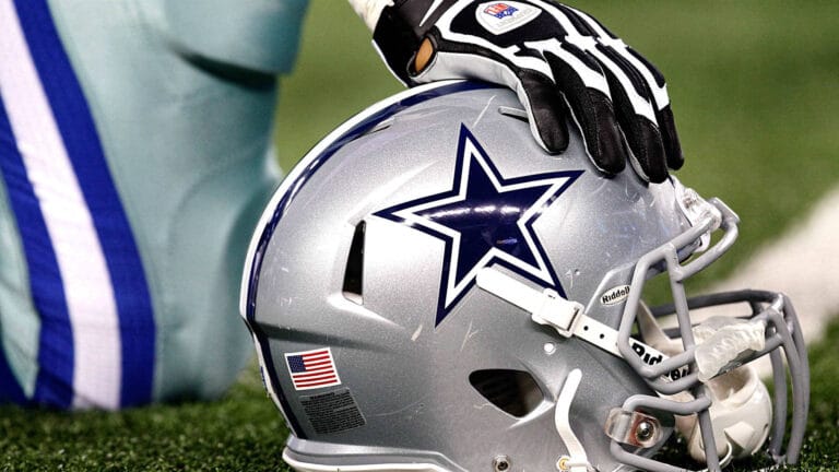 Football player resting a gloved hand on a Dallas Cowboys helmet lying on the turf.