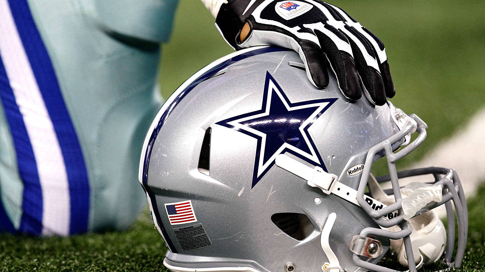 Football player resting a Dallas Cowboys helmet on the field, with a hand wearing a glove placed on top of the helmet.