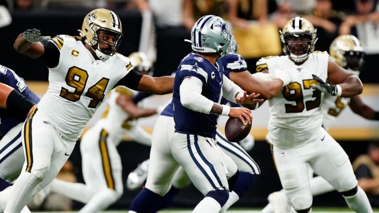 Football player in blue jersey evades two players in white jerseys on field during a game.