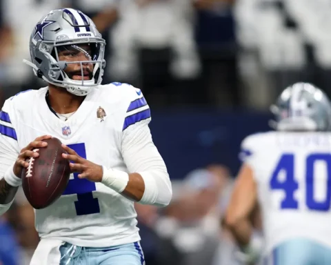 Football player in a white Dallas Cowboys uniform holding a football, preparing to throw during a game.