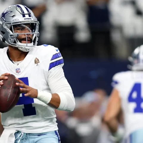 Football player in a white Dallas Cowboys uniform holding a football, preparing to throw during a game.