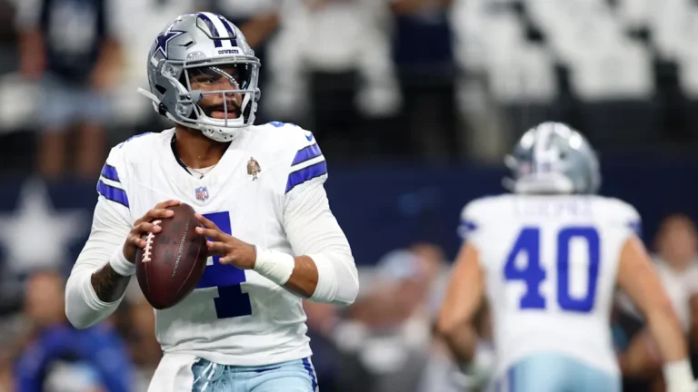 Football player in a white Dallas Cowboys uniform holding a football, preparing to throw during a game.