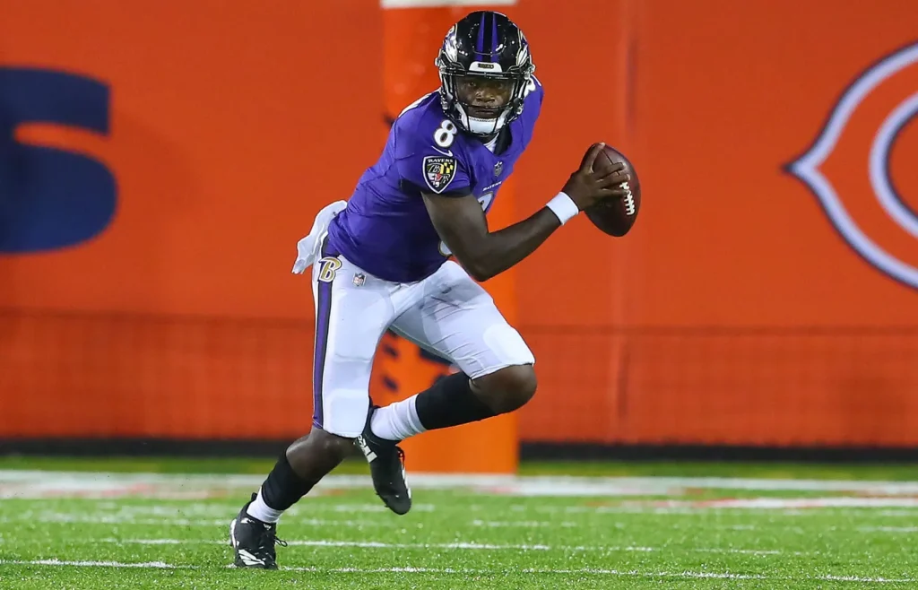 Football player in a purple jersey and white pants runs with the ball on an orange field.