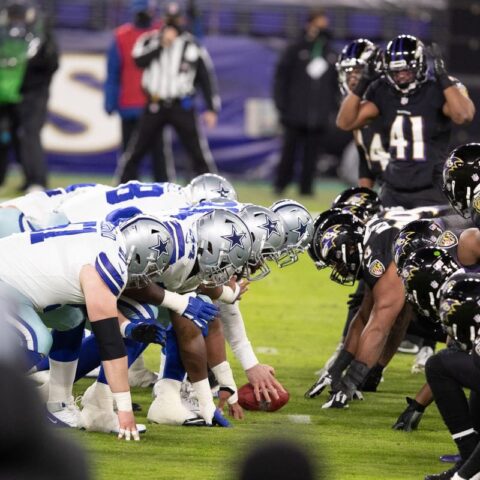 NFL teams line up for a snap, with the Cowboys in white uniforms and the Ravens in black uniforms, on a football field.