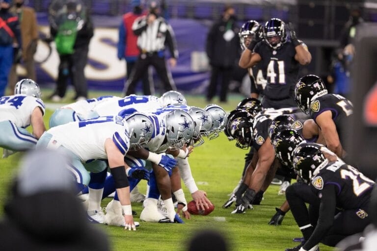 NFL teams line up for a snap, with the Cowboys in white uniforms and the Ravens in black uniforms, on a football field.