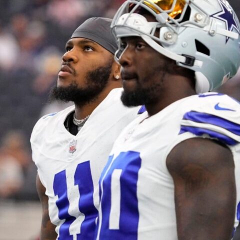 Two football players wearing white jerseys with dark blue numbers stand on the field, looking up.