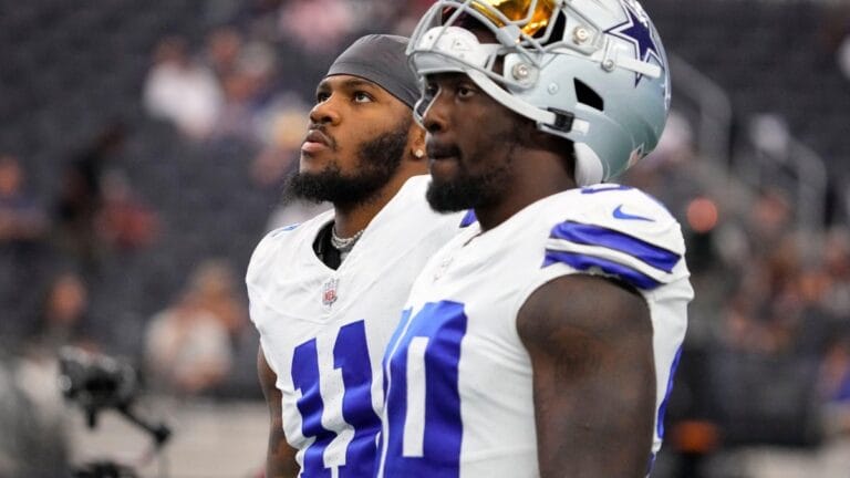 Two football players wearing white jerseys with dark blue numbers stand on the field, looking up.