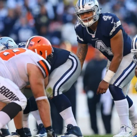 Football players from opposing teams line up for a play; a player in a blue uniform appears ready to receive the snap.