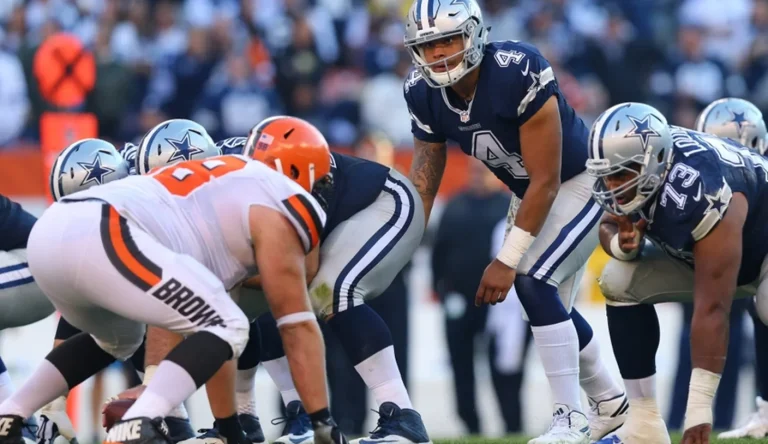 Football players from opposing teams line up for a play; a player in a blue uniform appears ready to receive the snap.