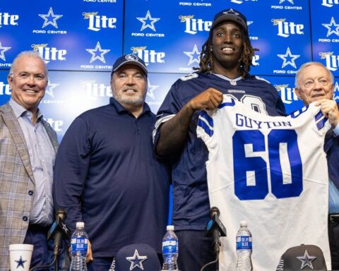 Four men stand together, with one holding up a "Glyton 60" football jersey, at a Dallas Cowboys press event backdrop.