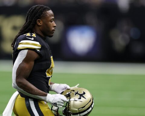 A player stands on the field holding his helmet, ready to contribute in his black and gold uniform.