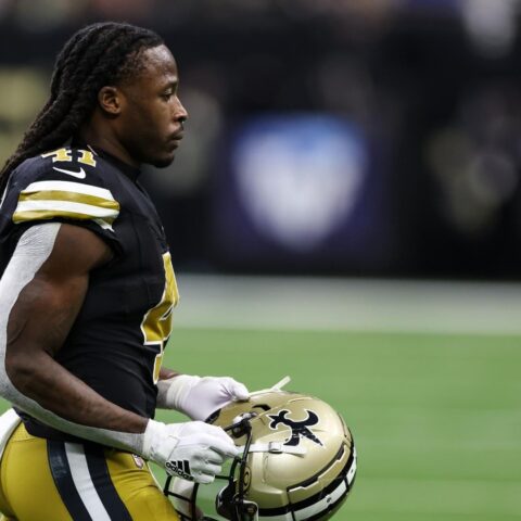 A player stands on the field holding his helmet, ready to contribute in his black and gold uniform.