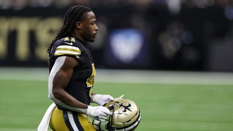 A player stands on the field holding his helmet, ready to contribute in his black and gold uniform.